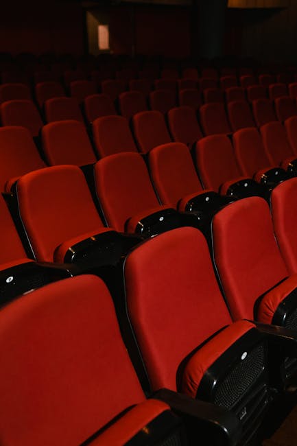 A wide-angle view of the auditorium at Fairfield Halls in Croydon, showing multiple rows of red upholstered seats with wooden armrests arranged on a sloped floor. The stage area, made of polished wood