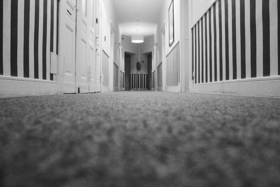 A low-angle view of a narrow, carpeted hallway in a residential home, showing a textured, neutral-colored carpet that appears clean and well-maintained. The walls are decorated with vertical striped w