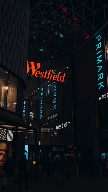 Interior view of the Westfield shopping mall in Croydon, showcasing a modern, multi-level space with high glass ceilings and structural steel supports illuminated with blue and red lighting. Prominent